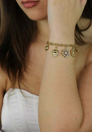 Vrouw draagt een gouden bedelarmband met zon-, bloem- en hartbedels om haar pols, gedeeltelijk haar gezicht bedekkend en een wit topje dragend.