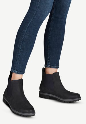 Botas negras hasta el tobillo con un acabado de cuero liso, paneles laterales elásticos y suelas de goma texturizada, combinadas con jeans ajustados oscuros.