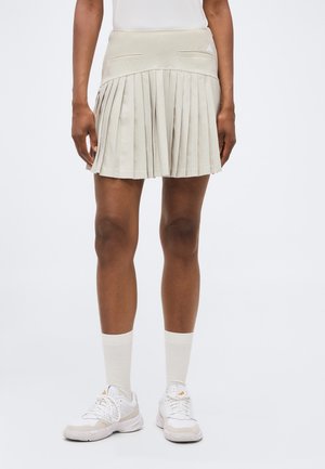 Kvinde iført beige plisseret nederdel, hvide sokker og hvide atletiksneakers, stående mod en ensartet lys baggrund.