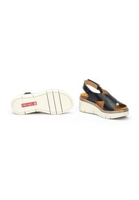 Sandalias de cuña de cuero negro con correas cruzadas, interior color tostado, suela blanca texturizada y un acento de relieve. Correa ajustable en el tobillo.