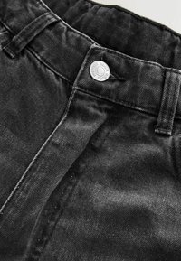 Gros plan sur la taille d'un jean en denim noir avec un bouton argenté portant l'inscription « Premium Denim Goods » et des passants de ceinture visibles.