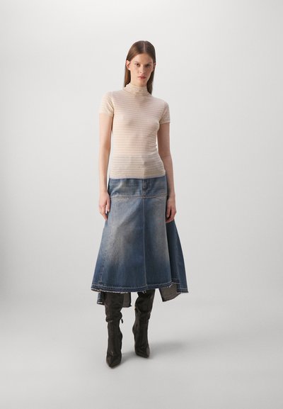 Bežs ribota adījuma turtlenecks tops, kopā ar garām džinsu svārkiem ar izplešanos apakšmalā un izbalējušu krāsu; modele nēsā melnus zābakus līdz ceļgaliem.
