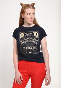 Námořnické tričko s designem Ouija desky v zlaté barvě, s kulatým výstřihem, krátkými rukávy a uvolněným střihem, kombinované s červenými kalhotami.