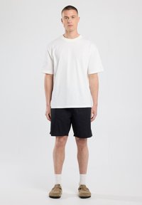 White cotton t-shirt with short sleeves, paired with black shorts. Worn with beige slip-on shoes and white crew socks. Simple design.