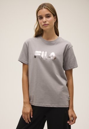Młoda kobieta z jasnobrązowymi włosami nosi szarą koszulkę FILA i czarne spodnie, stoi na tle jednolitego beżowego tła.