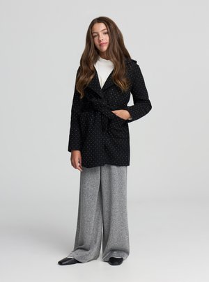 Cappotto nero a pois con cintura in vita, abbinato a pantaloni grigi a gamba larga con texture e scarpe nere con punta appuntita. La modella posa con sicurezza.