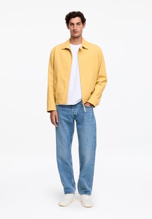 Mężczyzna stojący, ubrany w żółtą kurtkę na zamek, białą koszulkę, niebieskie dżinsy i białe trampki, na jednolitym białym tle.