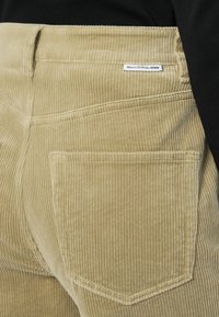 Lichtbeige corduroy broek met verticale ribbels, twee achterzakken en een klein merkembleem nabij de tailleband.