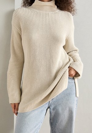 Chunky gebreide beige trui met een hoge geribbelde kraag, zijsplitten en lange mouwen. Zachte textuur en een relaxte pasvorm, gecombineerd met lichte spijkerbroek.