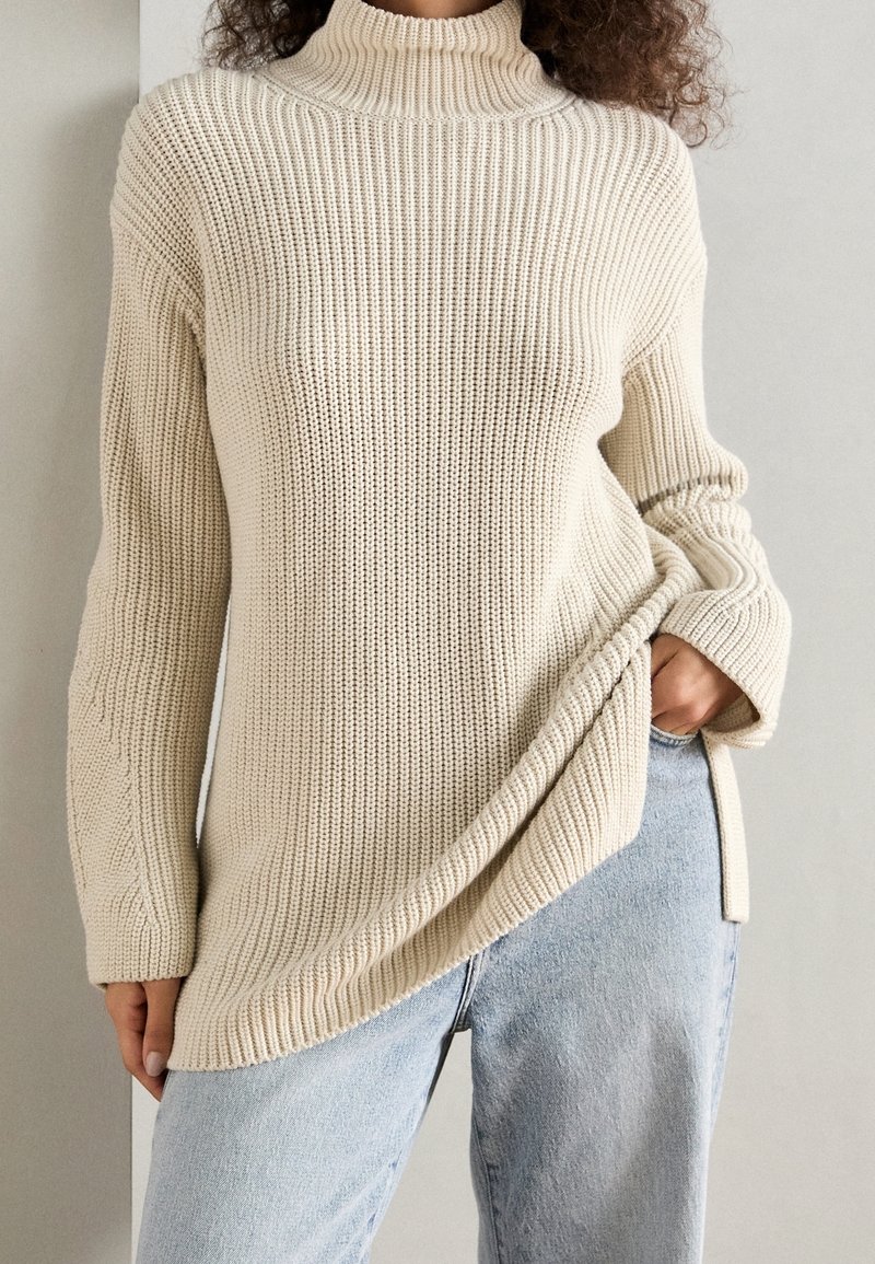 Chunky gebreide beige trui met een hoge geribbelde kraag, zijsplitten en lange mouwen. Zachte textuur en een relaxte pasvorm, gecombineerd met lichte spijkerbroek.