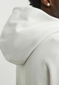 Lys grå hættetrøje med tekstureret stof, struktureret hætte og sømløst skulderdesign, der fremhæver minimalistiske detaljer.