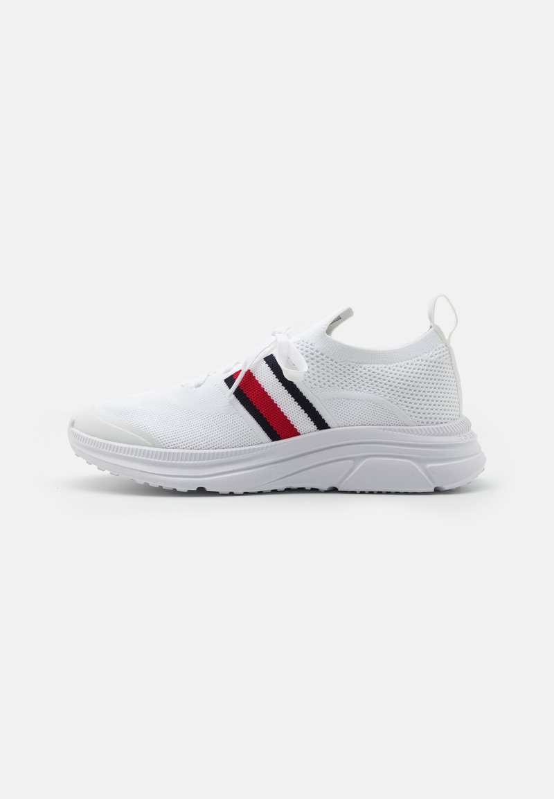 Bílé sportovní boty s prodyšným pleteným svrškem, s červenými a navy pruhy. Polstrovaná podešev s zaobleným tvarem a poutkem na patě.