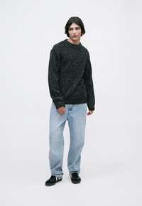 Tjock svart stickad tröja med rund halsringning, tillsammans med ljusblå löst sittande jeans och svarta sneakers. Enkla bakgrunden.