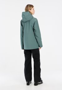 Wasserdichte grüne Jacke mit Kapuze, elastischen Bündchen und geradem Schnitt, kombiniert mit schwarzen Hosen und schwarzen Skischuhen. Glatte Textur, minimalistisches Design.