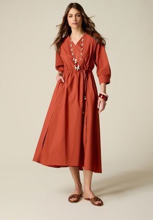 Femme portant une robe midi couleur rouille avec manches trois-quarts, collier en coquillage, bracelets et sandales plates marron, debout devant un fond uni.