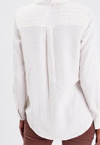 Chemise blanche à manches longues avec de fines rayures verticales et de petits points, dotée d'un ourlet arrondi et d'un pli au dos pour plus de mouvement.