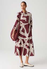 Abito camicia a motivi bordeaux e crema realizzato in tessuto leggero, con chiusura a bottoni frontale, vita arricciata e orlo a strati. Accompagnato da una borsa abbinata.
