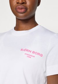 Valge puuvillane T-särk lühikeste varrukatega, mille vasakul rinnal on julge roosa tekst "BJÖRN BORG STOCKHOLM 1956". Sile tekstuur.