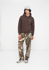 Joven vestido con una sudadera marrón, pantalones de camuflaje, zapatillas gris y blancas, y una gorra gris claro, de pie contra un fondo blanco.