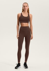 Smeđa sportska grudnjak i tajice od rastezljivog, tkanine koja upija vlagu. Moderan dizajn s visokim strukom i bez vidljivih šavova. Crne sportske tenisice.