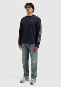 Marineblauwe longsleeve t-shirt met witte tekst op de linkermouw, gecombineerd met lichtblauwe versleten jeans en zilveren sneakers.