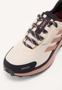 Lys beige treningssko med mesh og syntetisk overlegg, med mørke lisser, rosa detaljer, teksturert gummisåle, og «RAIN.RDY» etikett.