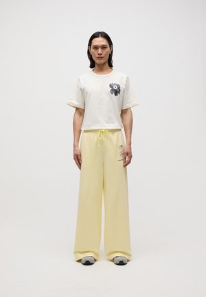 Pantaloni larghi di colore giallo chiaro con cordoncino e una t-shirt a maniche corte di colore crema con una grafica di fiore nera. Testo stampato sui pantaloni.