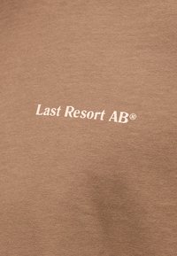 Țesătură bej cu text alb tipărit „Last Resort AB®” centrat pe suprafață.