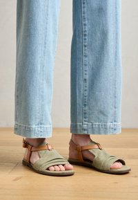 Sandalias de tela verde con detalles de cuero marrón, con la puntera descubierta y tiras entrelazadas, combinadas con unos vaqueros de pierna ancha de color azul claro.