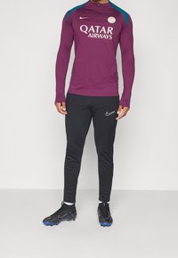 Fioletowa sportowa bluzka z długimi rękawami i niebieskimi akcentami, z wysokim kołnierzem i logo "Qatar Airways". Czarne spodnie dresowe Nike i czarne buty.