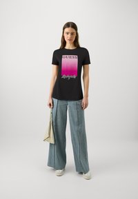 Zwarte T-shirt met een roze verloopprint met de tekst "GUESS" boven "Los Angeles," gecombineerd met lichtblauwe wijde broek en een beige tas.