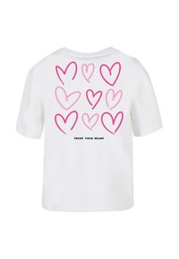 Weißes T-Shirt mit mehreren pinkfarbenen Herzdesigns auf der Rückseite, mit dem Text "VERTRAUE DEINEM HERZ" in Schwarz darunter gedruckt. Kurze Ärmel.