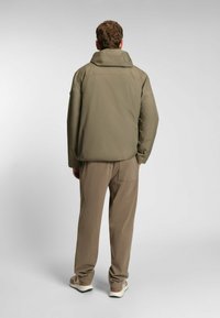 Giacca impermeabile verde oliva con colletto alto, grande cappuccio e maniche lunghe, abbinata a pantaloni marroni chiari e scarpe da ginnastica grigie.