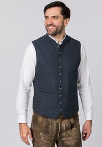 Marineblau gemusterte Weste mit sechs Metallknöpfen, einem hohen Kragen und zwei vorderen Taschen, getragen über einem weißen Hemd und braunen Hosen.