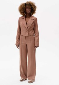 Bruine op maat gemaakte dubbele-breasted blazer met een cropped ontwerp, gecombineerd met wijde broekspijpen. Geaccessoreerd met geprinte schoenen en oorringen.
