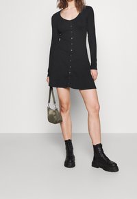 Robe longue noire côtelée à manches longues avec un décolleté arrondi et des boutons sur le devant, associée à des bottines noires à lacets et à un petit sac à main vert olive.