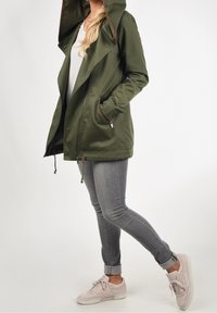 Oliivinvihreä hupullinen takki, jossa vetoketju, sivutaskut ja kiristysnyöri helmassa; yhdistetty harmaisiin kapeisiin farkkuihin ja vaaleanpunaisiin tennareihin.