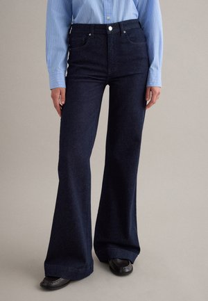 Persona che indossa jeans larghi blu scuro, camicia a righe blu e bianche infilata dentro, e scarpe nere, in piedi su uno sfondo neutro.