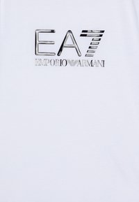 Camiseta blanca con un logo metálico plateado que dice "EA7 EMPORIO ARMANI". El material parece suave con un acabado ligeramente brillante.