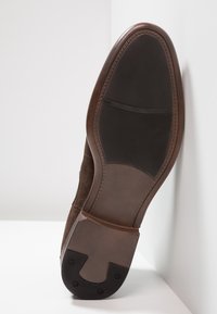 Semelle de chaussure en daim marron avec une semelle en caoutchouc noir texturé; comprend une forme incurvée et un talon arrondi, avec des détails de couture subtils.