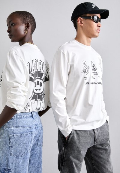 Des chemises blanches à manches longues présentent des impressions graphiques. Un porteur a un motif noir dans le dos ; l'autre porte un pantalon noir et des lunettes de soleil.