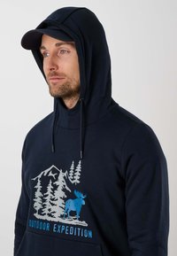 Homme portant un sweat à capuche bleu marine et une casquette, avec un graphique de montagnes, arbres et orignal bleu, texte "Outdoor Expedition" sur le devant.