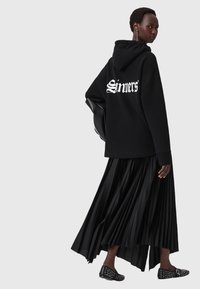Felpa nera oversize con la scritta "Sinners" sulla schiena, abbinata a una gonna nera plissettata e calzature nere con borchie.