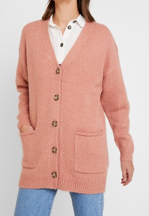 Kobieta ubrana w różowy dziergany sweter z dużymi guzikami i przednimi kieszeniami, na białej koszuli i niebieskich dżinsach, stojąca na jednolitym tle.