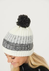 Vit stickad mössa med gråa och svarta ränder; har en stor svart pom-pom på toppen och ribbat mönster i basen.