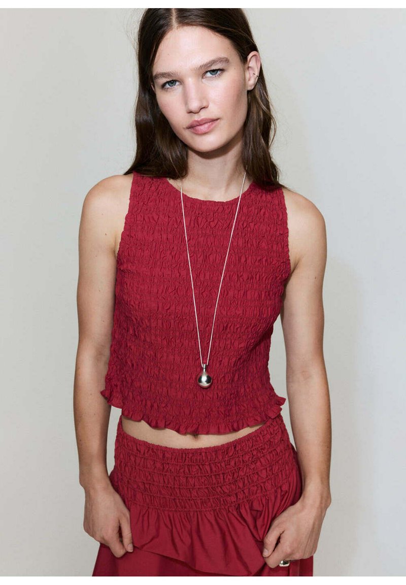 Jeune femme portant un crop top rouge texturé sans manches et une jupe assortie, avec un long collier argenté avec un pendentif rond.