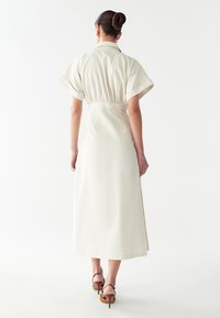 Abito crema, lungo fino al ginocchio, con maniche corte arrotolate, vita elasticizzata e silhouette dritta. Tessuto liscio con spacchi laterali.