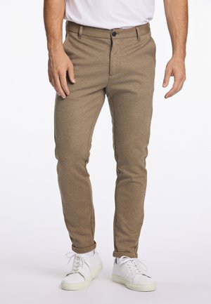 Mann trägt schlanke, beige Hose mit hochgekrempelten Säumen und weißen Turnschuhen, steht vor weißem Hintergrund.