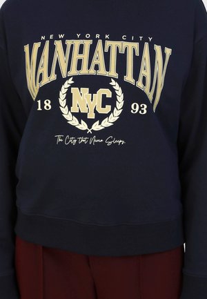 Námořnický svetr s výraznou bílou a zlatou grafikou, na které je napsáno "Manhattan," "NYC," a rok založení. Hladké manžety a lem.