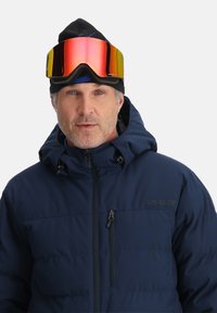 Marine blaue Pufferjacke mit hohem Kragen, Reißverschlusstaschen und matter Oberfläche, getragen mit einer schwarzen Mütze und reflektierenden Goggles.